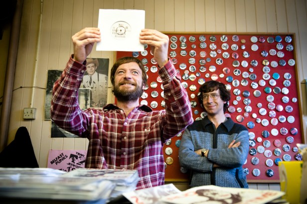 Terri Hooley (Richard Dormer) celebrates a Good Vibrations record release in the shop with Dave Hyndman (Michael Colgan). Hank Williams looks down benignly.